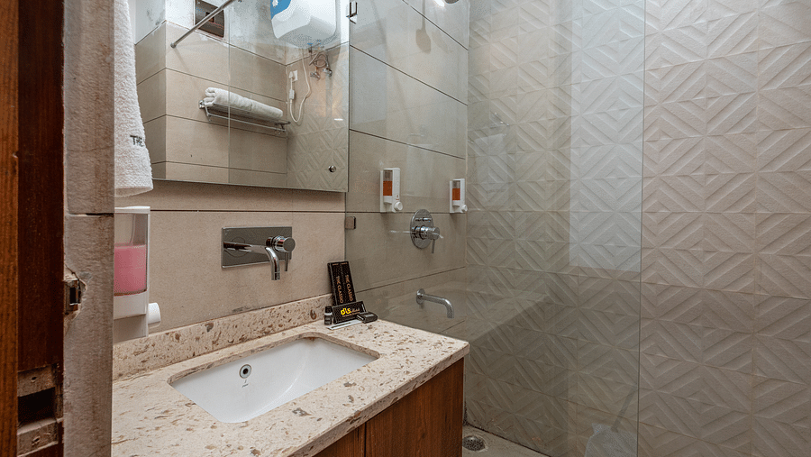 Side view of a bathroom showcasing a mirror above a washbasin with a wooden cabinet beneath and a shower space in the background at DLS The Classio